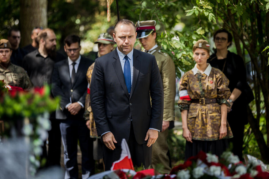 Mężczyzna w garniturze oddaje cześć nad grobem, jest w otoczeniu osób w mundurch