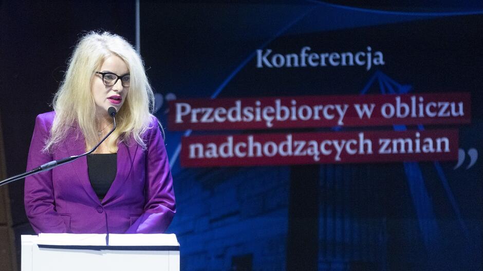 Na zdjęciu widać kobietę przemawiającą przy mównicy podczas konferencji. Kobieta ma długie blond włosy, nosi okulary i jest ubrana w elegancką, fioletową marynarkę oraz czarną bluzkę. Na tle znajduje się ciemnoniebieski baner z napisem w języku polskim: Konferencja Przedsiębiorcy w obliczu nadchodzących zmian , co sugeruje, że tematyka spotkania dotyczy wyzwań i zmian, przed którymi stoją przedsiębiorcy. Oświetlenie jest skoncentrowane na mównicy, co podkreśla centralną rolę mówczyni.