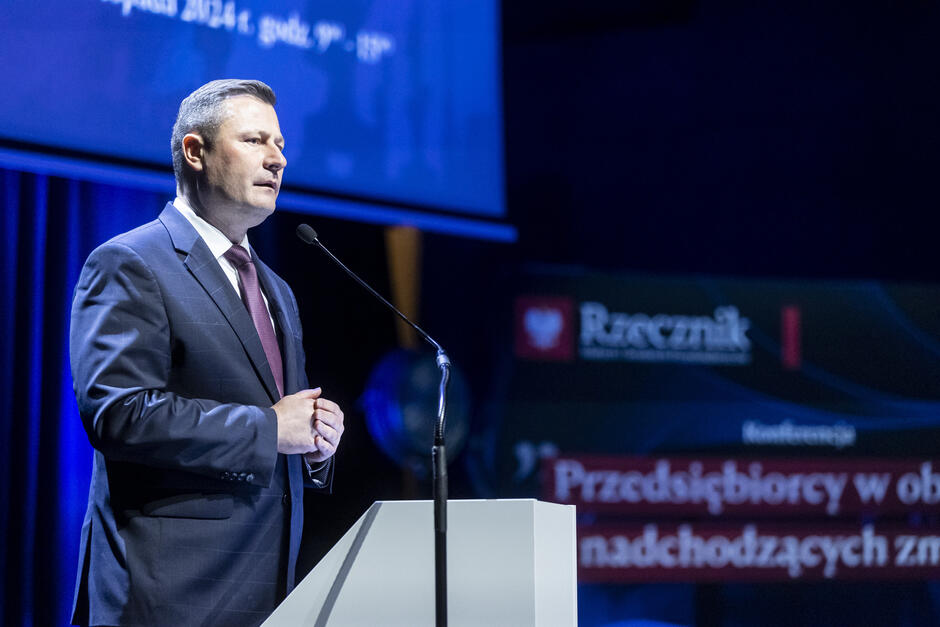 Na zdjęciu widzimy mężczyznę przemawiającego przy mównicy podczas oficjalnej konferencji. Mężczyzna jest ubrany w elegancki garnitur w ciemnym kolorze z delikatnym wzorem, białą koszulę i bordowy krawat. Ma krótkie, zaczesane włosy i stoi w profesjonalnej postawie, z rękami złożonymi na mównicy. W tle znajduje się ekran i baner z napisami, na którym widnieje logo i napis Rzecznik  oraz hasło konferencji:  Przedsiębiorcy w obliczu nadchodzących zmian . Oświetlenie jest skupione na mówcy, a tło pozostaje w półmroku, co nadaje scenie oficjalny i profesjonalny charakter.