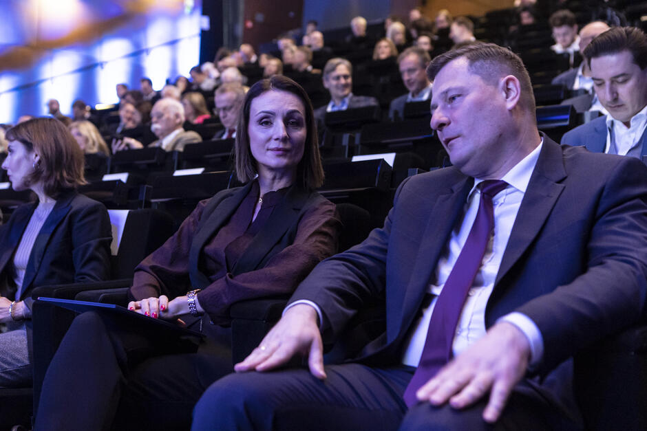 Zdjęcie przedstawia uczestników konferencji siedzących w sali audytoryjnej. Na pierwszym planie znajduje się kobieta w bordowej bluzce i ciemnej marynarce oraz mężczyzna w garniturze z bordowym krawatem. Kobieta patrzy w stronę kamery z lekkim uśmiechem, podczas gdy mężczyzna odwraca głowę w jej kierunku, jakby z nią rozmawiał. W tle widać innych uczestników konferencji siedzących w rzędach krzeseł, co tworzy atmosferę formalnego wydarzenia. Światło jest delikatne i niebieskawe, co nadaje scenie profesjonalny i nowoczesny charakter.