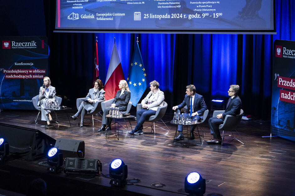 Zdjęcie przedstawia panel dyskusyjny odbywający się podczas konferencji w Europejskim Centrum Solidarności w Gdańsku. Na scenie siedzi sześć osób – cztery kobiety i dwóch mężczyzn – na nowoczesnych krzesłach, rozmieszczonych w półokręgu. Uczestnicy panelu są elegancko ubrani, co podkreśla formalny charakter wydarzenia. W tle widać flagi Polski, Unii Europejskiej i Gdańska, a także wyświetlony na ekranie program i szczegóły wydarzenia, m.in. data (25 listopada 2024 r.) oraz godziny (9:00–15:00). Po bokach sceny znajdują się banery promujące konferencję pod hasłem Przedsiębiorcy w obliczu nadchodzących zmian . Oświetlenie sceny skupia się na panelistach, natomiast sala jest przyciemniona, co tworzy profesjonalną atmosferę. Na stolikach przed uczestnikami widać karafki z wodą i szklanki, co podkreśla dbałość o komfort rozmówców.