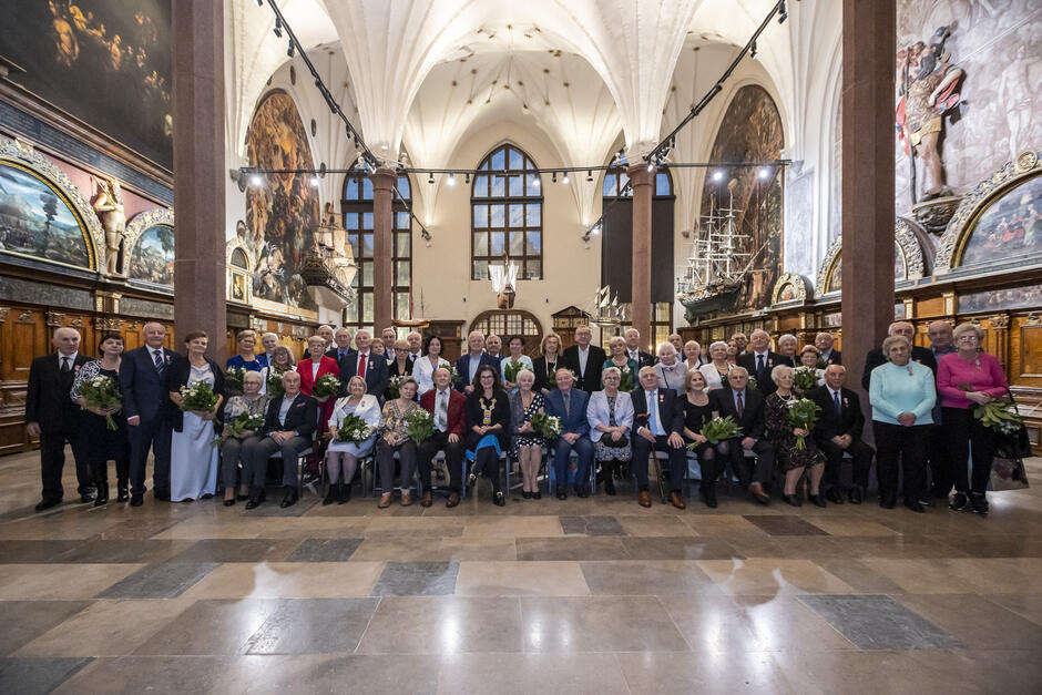 Na zdjęciu znajduje się grupa osób zgromadzona w eleganckim, zabytkowym wnętrzu, podczas uroczystości związanej z jubileuszami małżeńskimi. Tło wypełniają imponujące malowidła, modele statków i stylowe elementy architektoniczne, podkreślając podniosły charakter wydarzenia