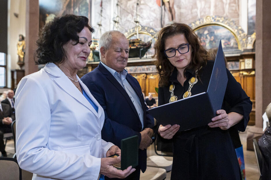 Na zdjęciu widać uśmiechniętą parę małżeńską podczas uroczystości, odbierającą pamiątkowe odznaczenie i dyplom od przedstawicielki władz, która przegląda dokument. W tle widoczne są zabytkowe dekoracje wnętrza z bogato zdobionymi elementami i modelami statków, podkreślając uroczysty charakter wydarzenia