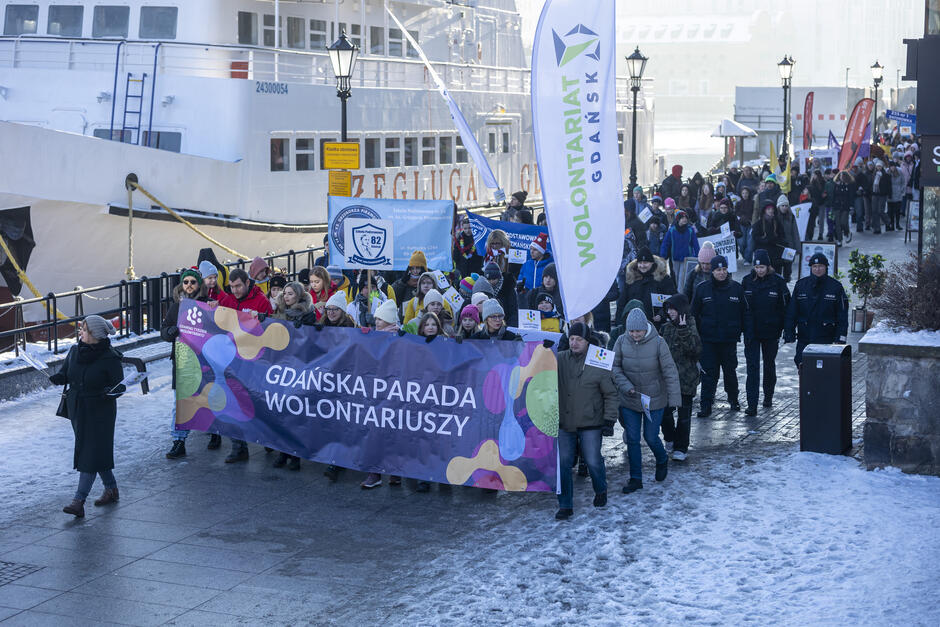 Zdjęcie przedstawia uczestników Gdańskiej Parady Wolontariuszy maszerujących wzdłuż nabrzeża, w tle widać statek z napisem Żegluga Gdańska . Grupa wolontariuszy niesie duży, kolorowy baner z napisem „Gdańska Parada Wolontariuszy” oraz flagi i transparenty z logotypami różnych organizacji i szkół, takich jak Szkoła Podstawowa nr 82. Uczestnicy są ciepło ubrani, co sugeruje zimową porę, dodatkowo potwierdzoną śniegiem widocznym na ziemi. W paradzie bierze udział wiele osób, zarówno młodszych, jak i starszych. Obok idą przedstawiciele organizatorów oraz funkcjonariusze służb miejskich. Wydarzenie przebiega w radosnej atmosferze, a zimowe słońce oświetla maszerujących, podkreślając wspólnotowy i aktywizujący charakter inicjatywy.