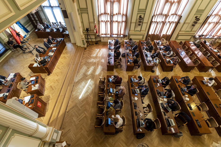 Zdjęcie przedstawia sesję w sali obrad rady miasta, w eleganckim wnętrzu z drewnianą podłogą i jasnymi zasłonami przepuszczającymi światło. Przy stołach siedzą radni, korzystając z laptopów, podczas gdy przestrzeń wypełnia atmosfera formalnej dyskusji i pracy.