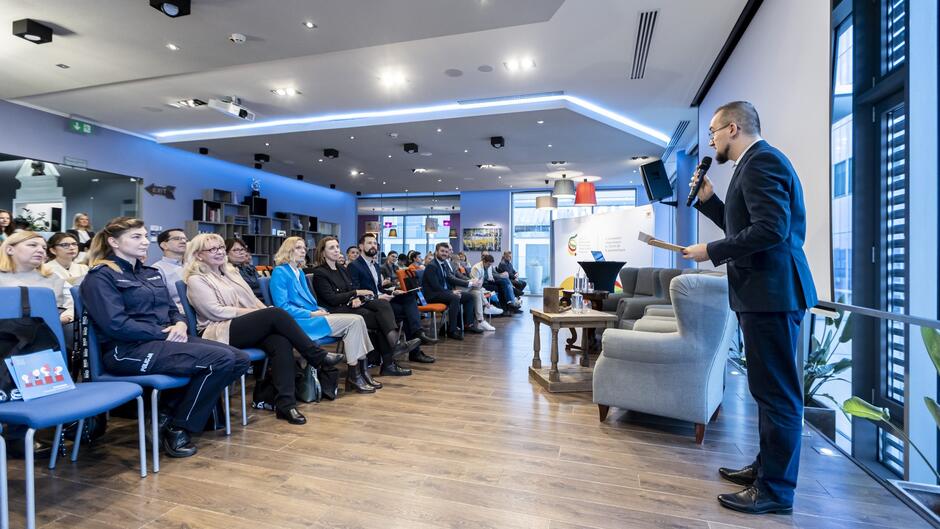 Zdjęcie przedstawia wydarzenie w formie spotkania lub konferencji odbywającej się w nowoczesnym, przestronnym wnętrzu. W pierwszym planie, po prawej stronie, znajduje się mężczyzna w garniturze, przemawiający do mikrofonu. Trzyma w ręku notatki, prowadząc prawdopodobnie prelekcję lub wprowadzając kolejne elementy programu. Na widowni zasiada różnorodna grupa osób, w tym przedstawiciele różnych środowisk, np. kobieta w mundurze policji, osoby w formalnych strojach biznesowych oraz inni uczestnicy. W tle widać jasne, nowoczesne oświetlenie, drewnianą podłogę i elementy wystroju wnętrza, takie jak regały z książkami czy ozdobne lampy. Za sceną można dostrzec stolik z wodą i notatkami, a także roll-up z logotypami i informacjami związanymi z wydarzeniem.  Atmosfera spotkania wydaje się formalna, ale jednocześnie przyjazna i otwarta na dialog.