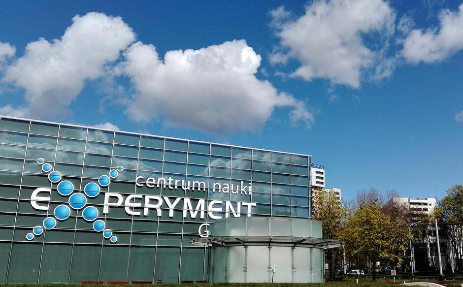 Na zdjęciu znajduje się Centrum Nauki Experyment w Gdyni, nowoczesny budynek z przeszkloną fasadą ozdobioną logo składającym się z niebieskich kul symbolizujących atomy. W tle widoczne są bloki mieszkalne, drzewa oraz błękitne niebo z białymi chmurami, co tworzy harmonijną i miejską atmosferę.