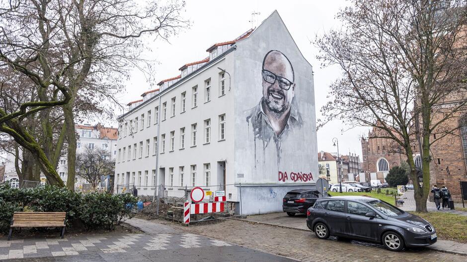 Zdjęcie przedstawia mural na ścianie budynku mieszkalnego w miejskim otoczeniu. Mural ukazuje portret uśmiechniętego mężczyzny z brodą i okularami, namalowany w realistycznym stylu. Pod portretem widnieje napis Dla Gdańska  w czerwonym kolorze. Budynek, na którym znajduje się mural, jest biały i ma charakterystyczne skośne dachy z czerwonymi dachówkami.  Na pierwszym planie widać wybrukowaną alejkę, ławkę oraz drzewa o bezlistnych gałęziach, co sugeruje, że zdjęcie wykonano w chłodniejszej porze roku. Po lewej stronie widoczna jest siatka ogrodzeniowa i znaki drogowe, które mogą wskazywać na trwające prace budowlane. Na ulicy zaparkowane są samochody, a w tle po prawej stronie można dostrzec fragment gotyckiego kościoła, co sugeruje, że scena rozgrywa się w historycznej części miasta.