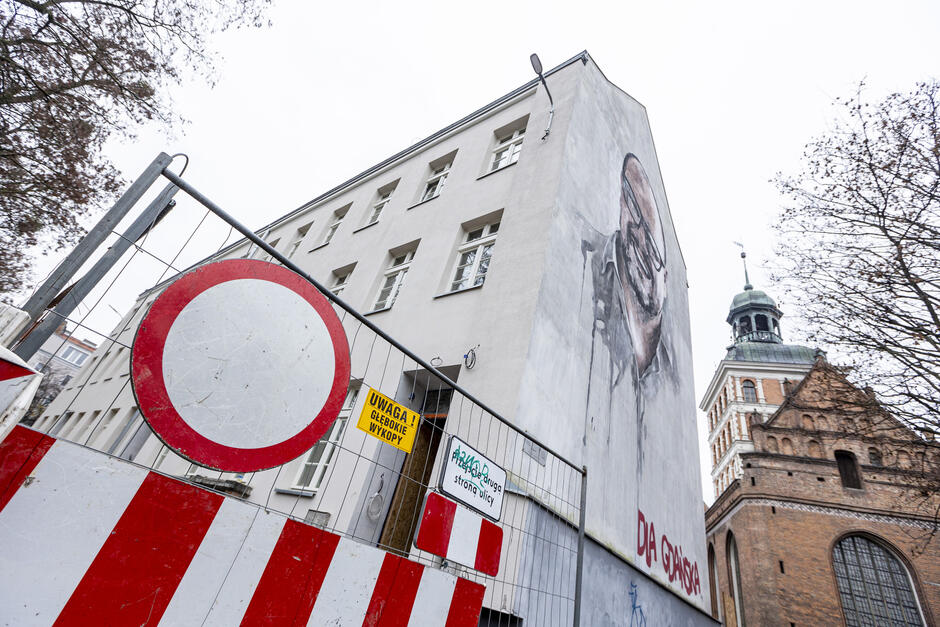 Zdjęcie ukazuje zbliżenie na budynek z muralem oraz fragment gotyckiego kościoła w tle. Na ścianie budynku widnieje realistyczny portret uśmiechniętego mężczyzny z napisem Dla Gdańska  w czerwonym kolorze.  Na pierwszym planie znajduje się ogrodzenie placu budowy, które zawiera znaki ostrzegawcze. Widać czerwono-biały znak zakazu wjazdu oraz żółtą tablicę z napisem  Uwaga! Głębokie wykopy . Jest też tabliczka informująca o przejściu na drugą stronę ulicy. Ogrodzenie podkreśla, że w miejscu prowadzone są prace budowlane.  Tło budynku zdobi wieża kościoła, która wyróżnia się swoim historycznym stylem i ceglaną architekturą. Ujęcie wykonane jest z niskiego kąta, co nadaje zdjęciu perspektywę i ukazuje szczegóły architektury oraz przestrzeń miejską.