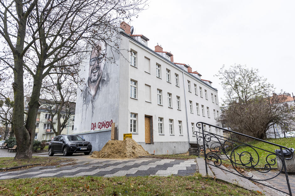 Zdjęcie przedstawia szerszą perspektywę budynku z muralem, na którym znajduje się portret uśmiechniętego mężczyzny z okularami oraz napis Dla Gdańska  w czerwonym kolorze. Budynek jest biały, wielokondygnacyjny, z czerwonym dachem. Na dole ściany widać otwór drzwiowy zabezpieczony płytą, a przed nim znajduje się stos piachu, co wskazuje na trwające prace remontowe lub budowlane.  Na pierwszym planie widoczna jest ozdobna balustrada schodów prowadzących do chodnika oraz fragment zieleni z trawą. Po lewej stronie widać drzewo bez liści, co sugeruje zimową lub późnojesienną porę roku. W tle znajdują się inne budynki mieszkalne, a po lewej zaparkowany jest ciemny samochód. Całość ukazuje miejską scenerię w otoczeniu zabytkowej i mieszkalnej architektury.