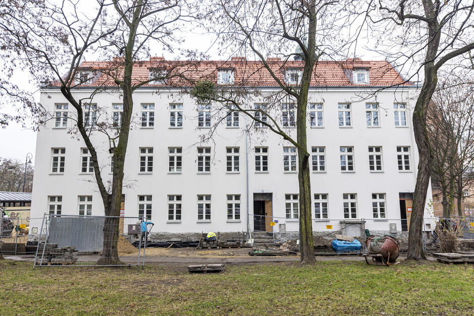 Zdjęcie ukazuje frontową elewację białego, czterokondygnacyjnego budynku z czerwonym dachem w trakcie prac budowlanych lub remontowych. Wzdłuż elewacji ustawione są ogrodzenia budowlane, za którymi widać stosy materiałów, takie jak deski, gruz i narzędzia. Na ziemi leżą także elementy budowlane, m.in. palety, worki z materiałami oraz betoniarka. Przy budynku pracują robotnicy ubrani w odzież ochronną. Na pierwszym planie widoczny jest trawnik z drzewami, które mają gołe gałęzie, co wskazuje na późnojesienną lub zimową porę. W tle widać niewielkie fragmenty innych obiektów miejskich, w tym budynków oraz kościoła, którego wieża jest widoczna za drzewami po prawej stronie. Sceneria przedstawia spokojną miejską okolicę w trakcie remontu infrastruktury.