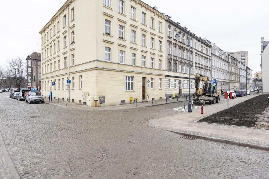 na zdjęciu fragment brukowanej ulicy, widać też nowe chodniki, w tle kamienice, a przed nimi żółta koparka i kilka zaparkowanych samochodów