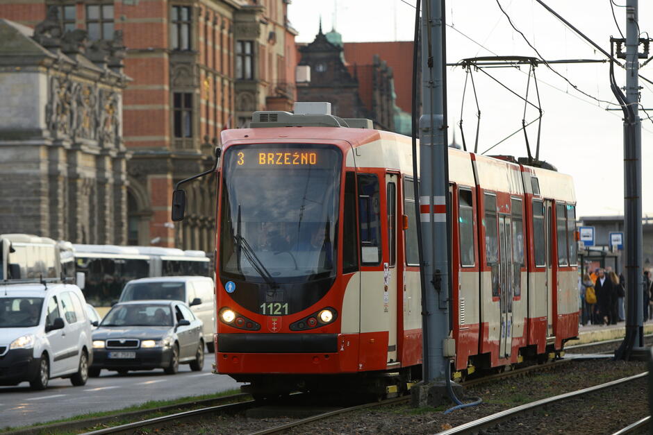 Na zdjęciu widać czerwono-kremowy tramwaj o numerze 1121 z napisem 3 Brzeźno  na wyświetlaczu, jadący przez miasto w otoczeniu zabytkowej architektury i ruchu ulicznego. W tle widoczne są charakterystyczne budynki i ludzie czekający na przystanku