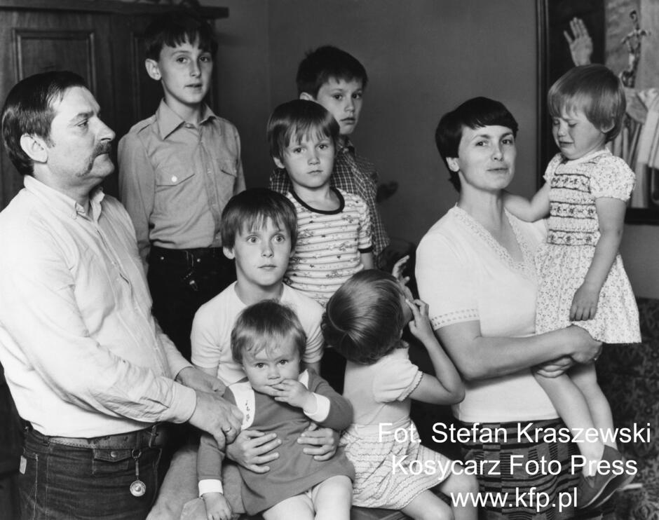 Czarnobiałe zdjęcie wielodzietnej rodziny. Po lewej stronie kadr zamyka mężczyzna z dużymi wąsami po prawej widać krótko obciętą kobietę trzymającą na rękach małą dziewczynkę