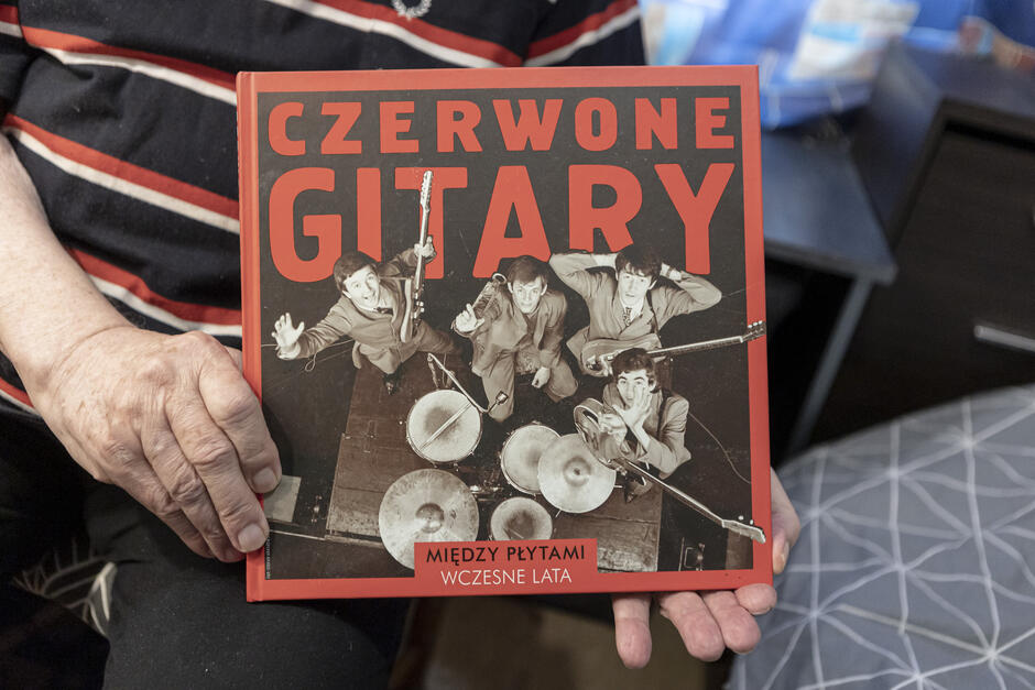 kolorowe zdjęcie przedstawia ręce trzymające okładkę książki z widocznym na niej czarnobiałym zdjęciem zespołu muzycznego stojącego przy perkusji