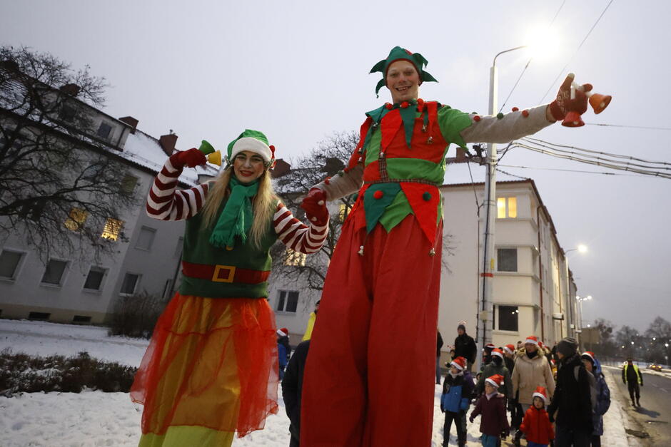 parada przebierańców, ubrany kolorowo, z przewagą zielonego i czerwonego skrzat i mikołajka na szczudłach, w tle białe budynki