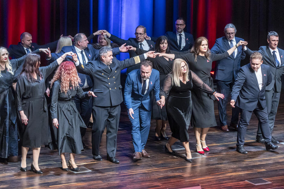 Na zdjęciu widzimy grupę dorosłych osób uczestniczących w tańcu na scenie. Osoby trzymają się za ręce, tworząc złączone ramiona i podniesione dłonie, co może sugerować tradycyjny polski taniec, np. poloneza. Uczestnicy są elegancko ubrani – kobiety mają czarne sukienki, a mężczyźni garnitury, jeden z nich ubrany jest w mundur marynarski. Na piersiach wielu osób widać biało-czerwone kotyliony, co wskazuje, że wydarzenie ma charakter patriotyczny, prawdopodobnie związany z obchodami święta narodowego. Tło sceny jest podświetlone na biało-czerwono, co dodatkowo podkreśla narodowy charakter uroczystości. Atmosfera zdjęcia jest uroczysta i pełna zaangażowania.