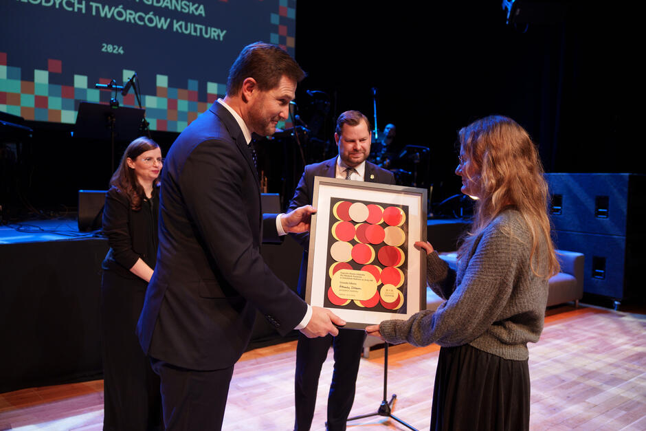 Na zdjęciu widzimy moment wręczenia nagrody młodemu twórcy kultury podczas uroczystej gali. Osoba wręczająca dyplom jest elegancko ubrana, a w tle znajduje się scena z oświetleniem, co podkreśla oficjalny charakter wydarzenia