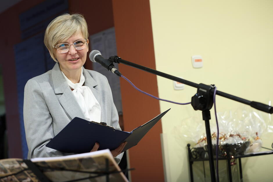 Kobieta po pięćdziesiątce, z krótkimi blond włosami, w okularach. Jest ubrana w szarą garsonkę i szare spodnie do kompletu