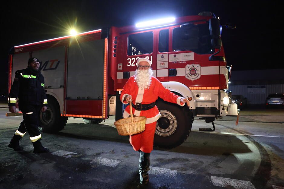 Na zdjęciu widoczny jest mężczyzna przebrany za Świętego Mikołaja, który niesie koszyk, a w tle stoi strażacki wóz bojowy OSP z włączonymi światłami. Obok Mikołaja znajduje się strażak, co sugeruje udział w świątecznej akcji organizowanej przez straż pożarną.