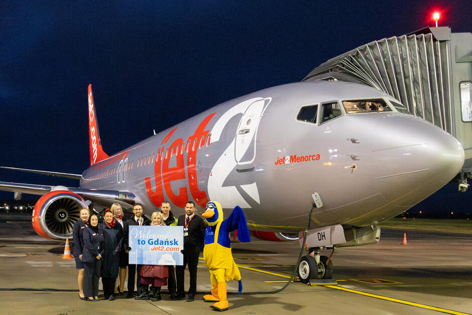 Zdjęcie przedstawia samolot linii lotniczej Jet2, stojący na płycie lotniska, z napisem „Jet2Menorca” widocznym na jego kadłubie. Przed samolotem grupa pracowników oraz maskotka w kostiumie ptaka trzymają tablicę z napisem „Welcome to Gdańsk” na tle nocnego nieba.