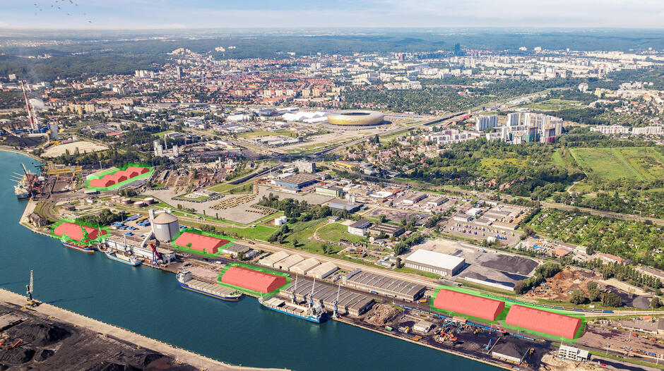 Zdjęcie przedstawia panoramiczny widok portu morskiego i jego okolic, wykonane z lotu ptaka. Na pierwszym planie widoczny jest nabrzeże portowe z zacumowanymi statkami, magazynami i dźwigami. Niektóre budynki i obszary na terenie portu zostały oznaczone na czerwono i zielono, co może wskazywać na ich szczególne przeznaczenie lub etapy planowania inwestycji. W tle rozciąga się miasto z licznymi budynkami mieszkalnymi, biurowymi oraz stadionem o charakterystycznym, złotym kształcie. Z prawej strony widoczne są tereny zielone i ogródki działkowe. Po lewej stronie znajduje się infrastruktura przemysłowa, w tym kominy fabryczne oraz place przeładunkowe.  Całość zdjęcia pokazuje zróżnicowany krajobraz łączący tereny przemysłowe, portowe i miejskie. Perspektywa z góry umożliwia uchwycenie skali i organizacji przestrzennej tego obszaru.