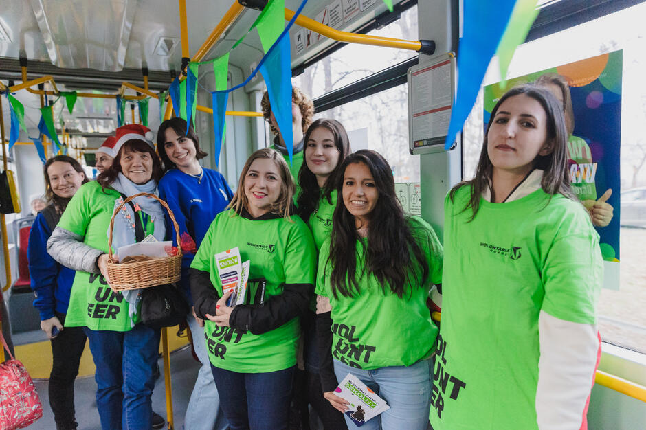 Na zdjęciu znajduje się grupa wolontariuszy w tramwaju, ubranych w zielone koszulki z napisem Wolontariat Banku . Wszyscy są uśmiechnięci, trzymają broszury i kosz z upominkami, a wnętrze tramwaju jest ozdobione kolorowymi girlandami, co wskazuje na akcję charytatywną lub promocyjną.