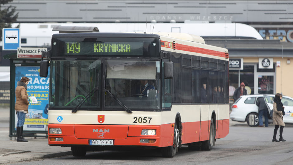 Na zdjęciu widzimy autobus marki MAN, należący do komunikacji miejskiej w Gdańsku. Pojazd ma numer boczny 2057 i obsługuje linię 149, której kierunek wskazany na wyświetlaczu to Krynicka . Autobus jest w barwach typowych dla gdańskiej komunikacji miejskiej – kremowo-czerwonych. W tle widać przystanek autobusowy  Wrzeszcz PKP  z tabliczką przystankową oraz budynek dworca kolejowego w dzielnicy Wrzeszcz. W pobliżu autobusu znajdują się przechodnie – jedna osoba czeka na przystanku, a inna przechodzi obok. Scena ma charakter codzienny i miejskiego ruchu.