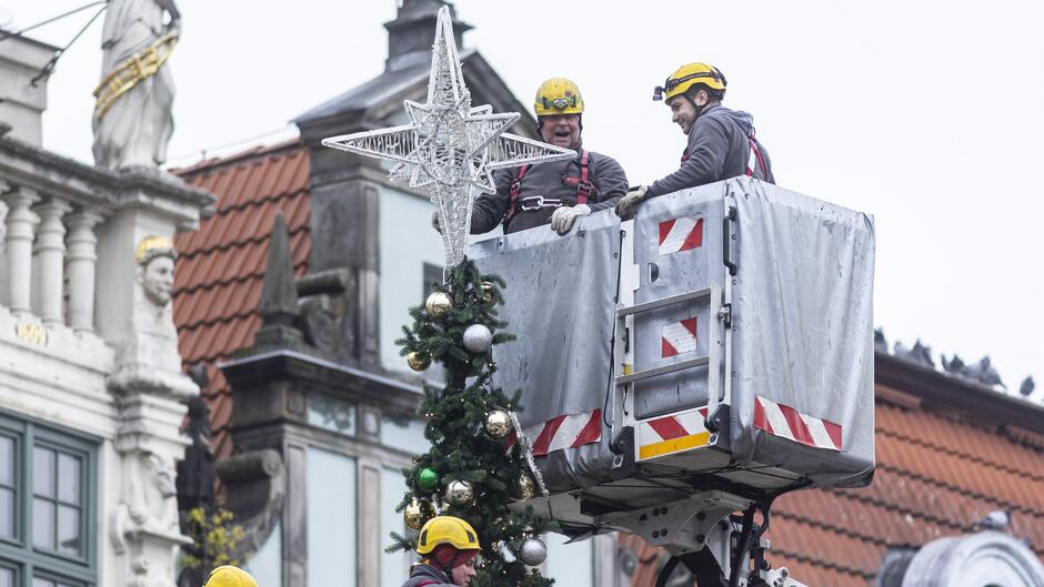 Na zdjęciu widzimy pracowników zajmujących się montażem świątecznej choinki na tle zabytkowej architektury. Pracownicy znajdują się w podnośniku koszowym, a jeden z nich instaluje gwiazdę na szczycie choinki. Drzewko jest ozdobione bombkami w kolorach złotym, srebrnym i zielonym, co dodaje świątecznego charakteru. W tle widoczne są budynki z czerwonymi dachówkami i zdobieniami, charakterystyczne dla historycznej zabudowy, najprawdopodobniej na Długim Targu w Gdańsku. Scena przedstawia przygotowania do nadchodzących świąt Bożego Narodzenia.