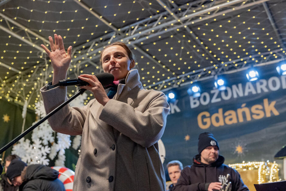 Zdjęcie przedstawia kobietę przemawiającą na scenie podczas wydarzenia świątecznego w Gdańsku, ozdobionego światełkami i dekoracjami w tle, takimi jak śnieżynki. W tle widać również innych uczestników wydarzenia oraz napis Gdańsk  na banerze