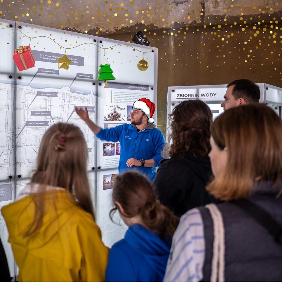 Zdjęcie przedstawia grupę osób uczestniczących w edukacyjnym oprowadzaniu, prawdopodobnie w muzeum lub na wystawie związanej z historią infrastruktury wodnej. Przewodnik ubrany w niebieską bluzę i czapkę Mikołaja wyjaśnia coś na tle plansz informacyjnych, które przedstawiają mapy, schematy lub zdjęcia związane z tematyką zbiorników wody lub infrastruktury. Na górze plansz znajdują się świąteczne ozdoby graficzne, takie jak bombki, choinka oraz prezenty, które wprowadzają wesoły, świąteczny nastrój do naukowej i edukacyjnej atmosfery wydarzenia. Grupa słuchaczy wygląda na zaangażowaną w słuchanie, co wskazuje na interesujące i dobrze prowadzone oprowadzanie.