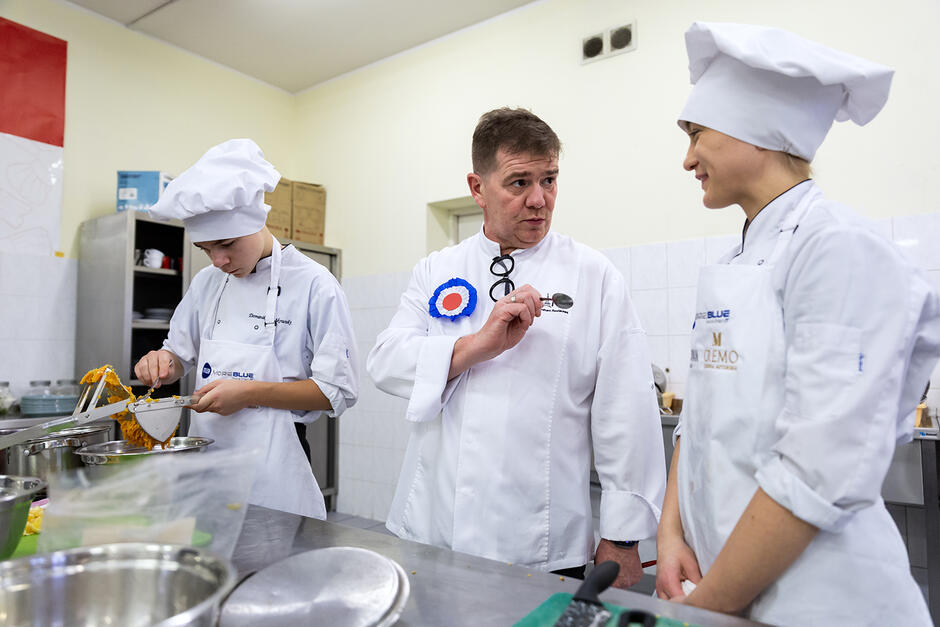 Klasa szkoły gastronomicznej. Kucharz mówi do uczennicy w stroju kucharskim.