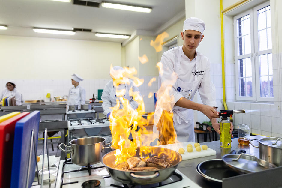 Uczeń szkoły gastronomicznej w stroju kucharskim gotuje w klasie, na patelni wybucha kula ognia.