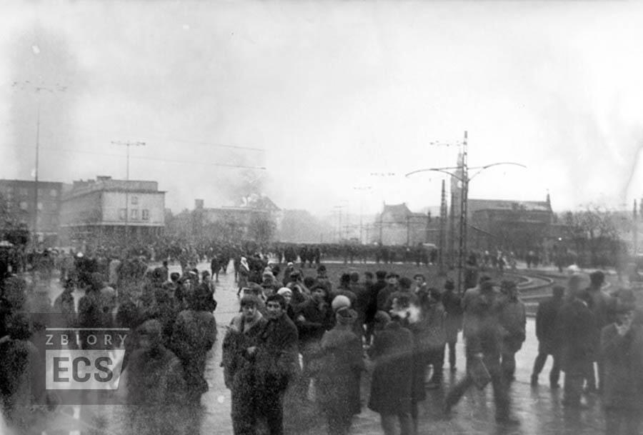 Zdjęcie przedstawia scenę z protestów w Gdańsku w grudniu 1970 roku, związanych z rewolucją grudniową. Widać tłum ludzi zgromadzonych na ulicy w napiętej atmosferze, a częściowo zamglone tło oddaje chaos tamtych dramatycznych wydarzeń