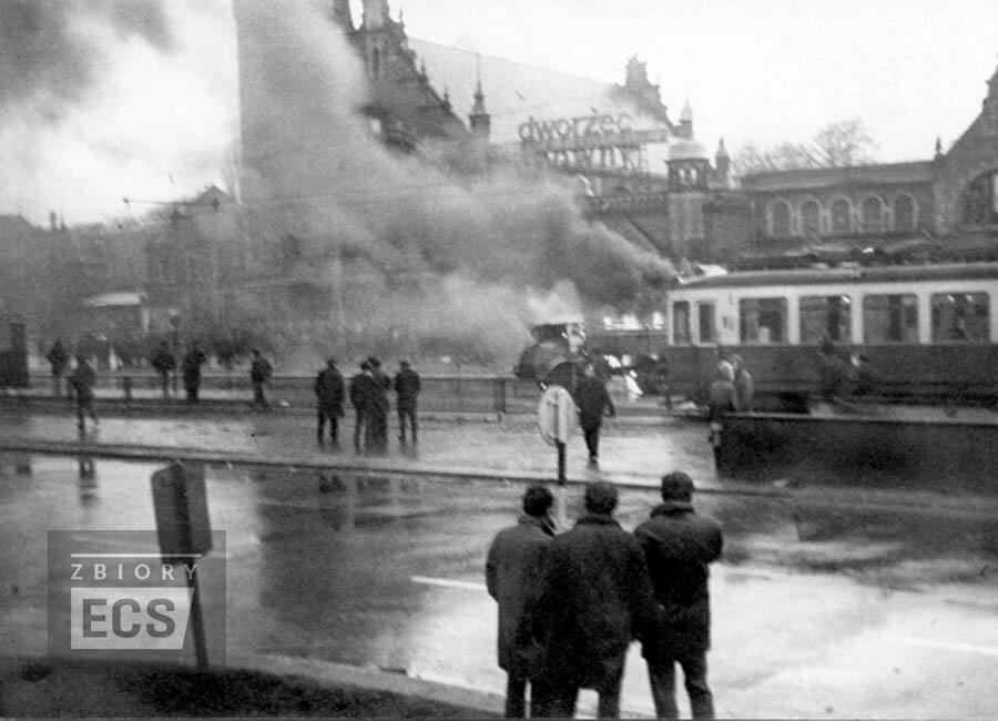 Zdjęcie przedstawia dramatyczną scenę z protestów w Gdańsku w grudniu 1970 roku, z widocznym płonącym pojazdem i gęstym dymem unoszącym się w okolicy dworca głównego. Na pierwszym planie widać kilka osób obserwujących zdarzenie, co podkreśla napiętą i chaotyczną atmosferę tych wydarzeń