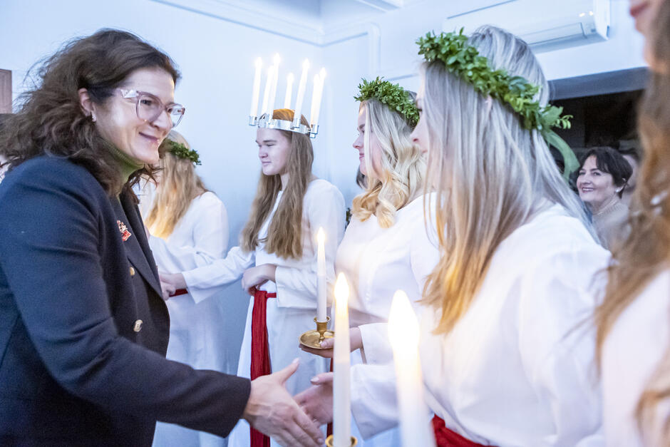 Dziewczyny w białych strojach i wiankach na głowach, jedna ma na głowie świece, witają się z kobieta - prezydent Gdańska.