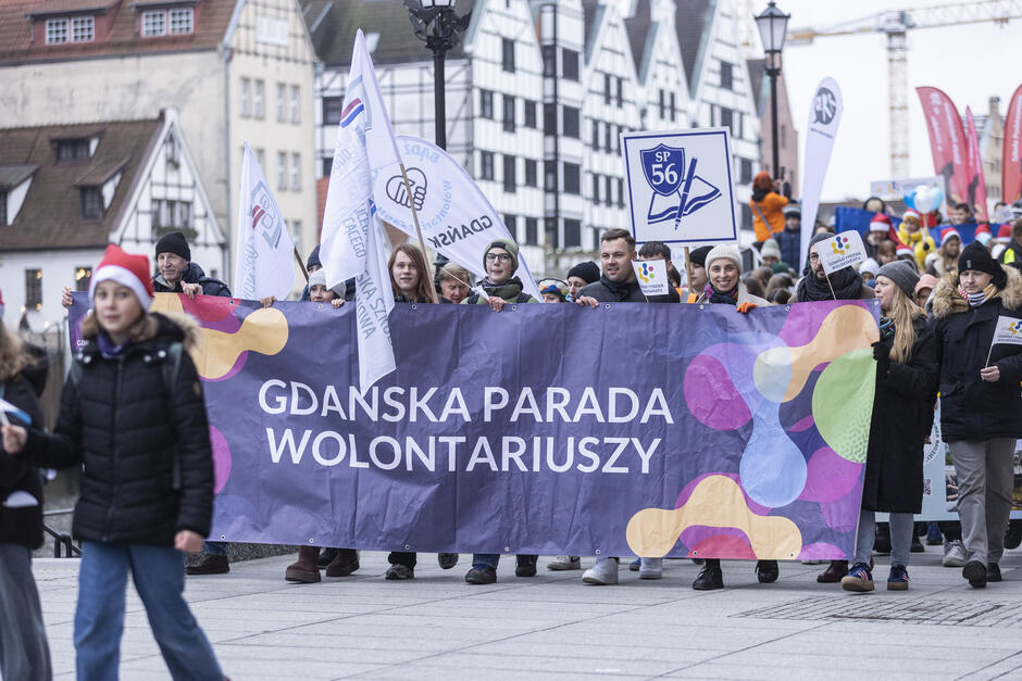 na zdjęciu barwna parada, na czele idzie kilka osób z podłużnym plakatem z napisem parada wolontariuszy