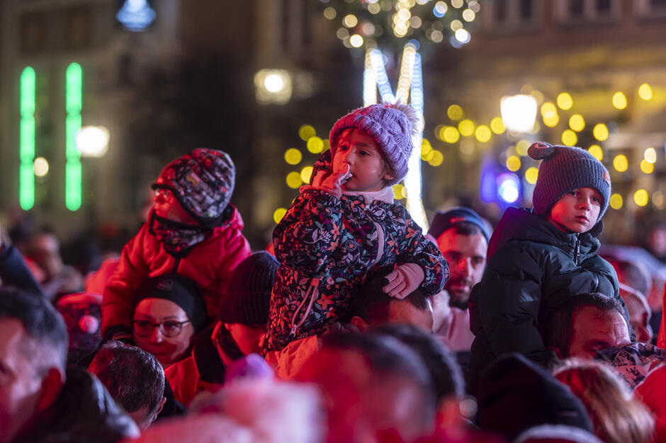 Na zdjęciu widzimy grupę ludzi zgromadzonych podczas wieczornego wydarzenia, oświetlonych ciepłym, świątecznym światłem. Na pierwszym planie dzieci siedzą na ramionach dorosłych, otulone w zimowe kurtki i czapki, z wyrazem zainteresowania lub zadumy na twarzach, a w tle migoczą świąteczne dekoracje.