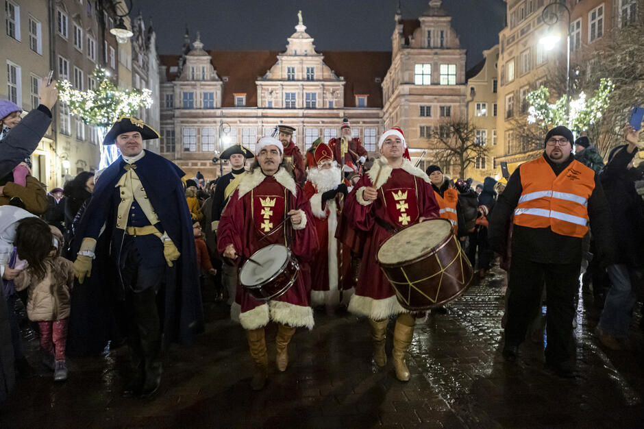 Na zdjęciu przedstawiono paradę na tle zabytkowych kamienic, z udziałem osób ubranych w stroje historyczne i świąteczne. Na pierwszym planie widoczni są bębniarze w czerwonych płaszczach z herbem oraz postać Świętego Mikołaja, a wokół gromadzi się tłum obserwatorów w zimowej scenerii.