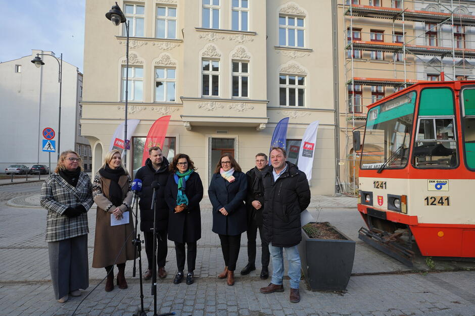 Na zdjęciu znajduje się grupa osób stojących przed odnowioną kamienicą oraz tramwajem, symbolizującym lokalną komunikację miejską. Ustawione flagi oraz mikrofony sugerują, że odbywa się oficjalne wydarzenie, być może związane z zakończeniem renowacji lub prezentacją projektu.