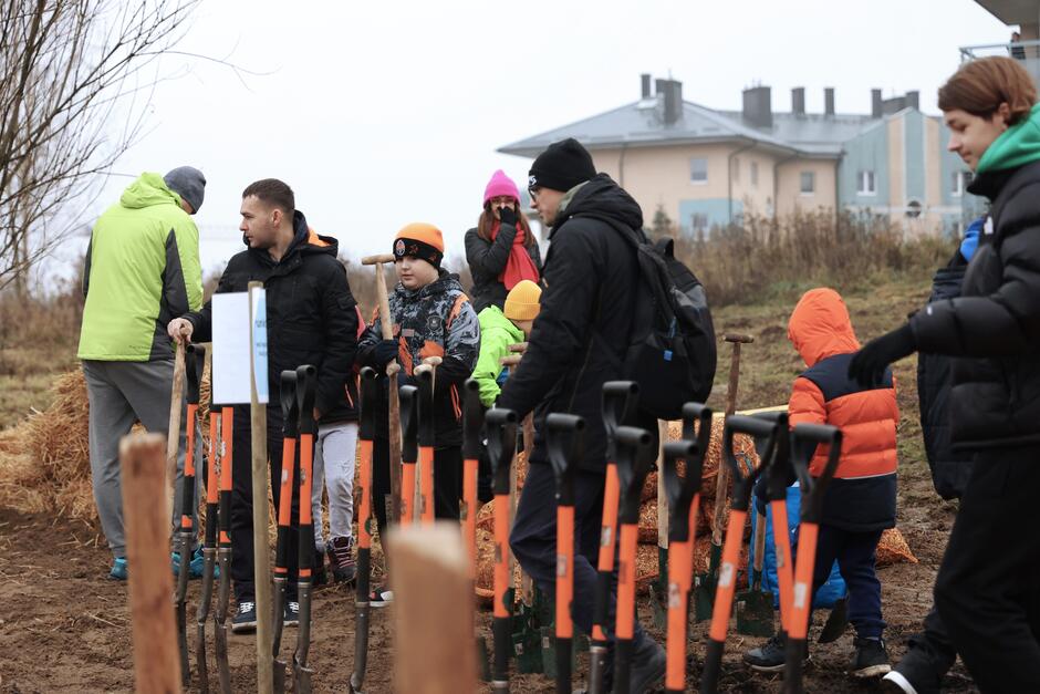 grupa osób w ciepłych kurtkach, na pierwszym planie łopaty wbite w ziemię