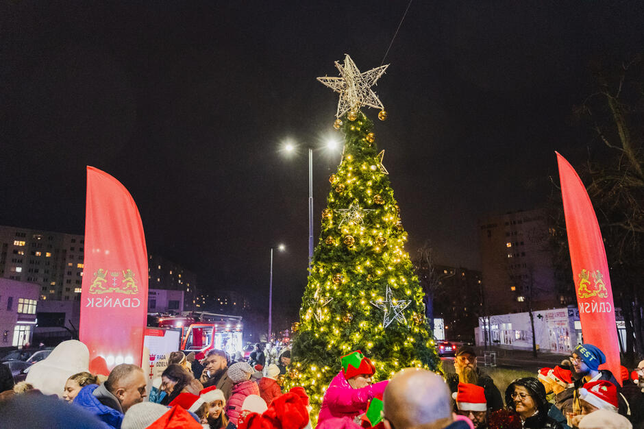 na zdjęciu rozświetlona choinka, wokół niej tłum ludzi w zimowych strojach