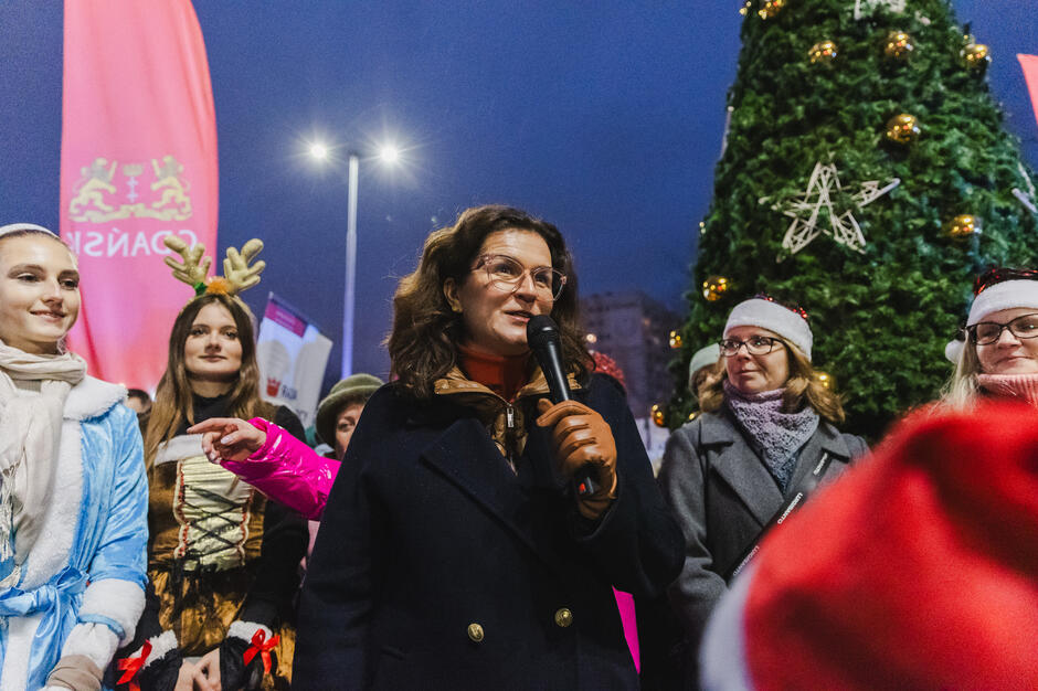 na zdjęciu kobieta w ciemnym płaszczu, to prezydent gdańska, mówi do mikrofonu, wokół niej stoi kilka osób w zimowych strojach, niektóre w bajkowych kostiumach