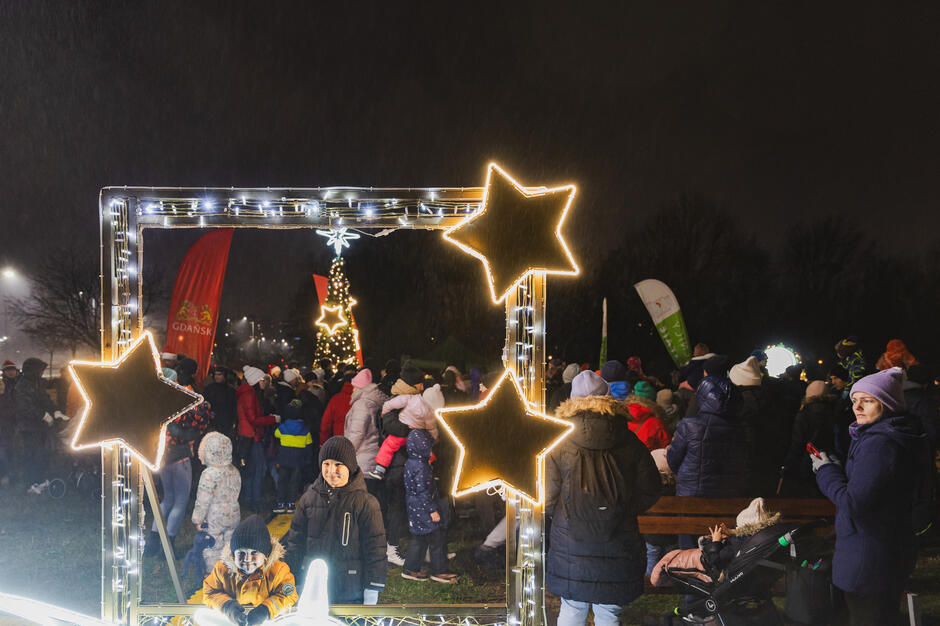 na pierwszym planie iluminacja przypominająca ramę obrazu z gwiazdami, w tle tłum ludzi i rozświetlona choinka