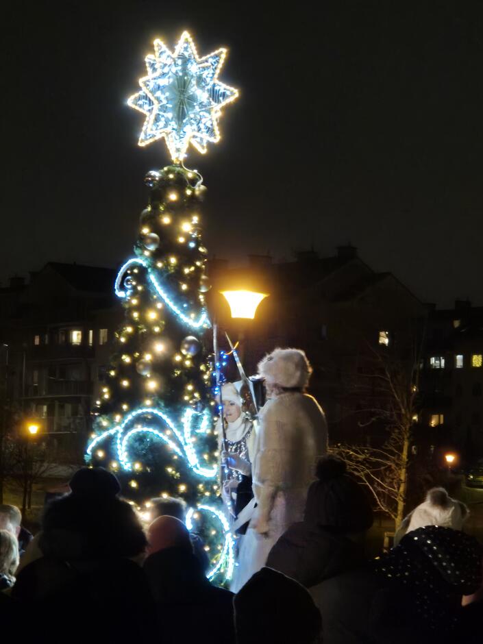 choinka i kobieta na szczudłach w białym kostiumie, obok grupka ludzi