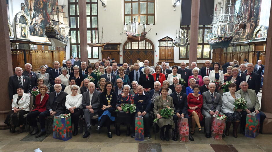 Zdjęcie przedstawia dużą grupę osób, głównie seniorów, zgromadzonych w eleganckim wnętrzu z wysokimi oknami, modelami żaglowców i bogato zdobionymi detalami architektonicznymi. W centralnej części siedzą odznaczone pary, które trzymają bukiety kwiatów, co sugeruje uroczystość jubileuszową, taką jak złote gody