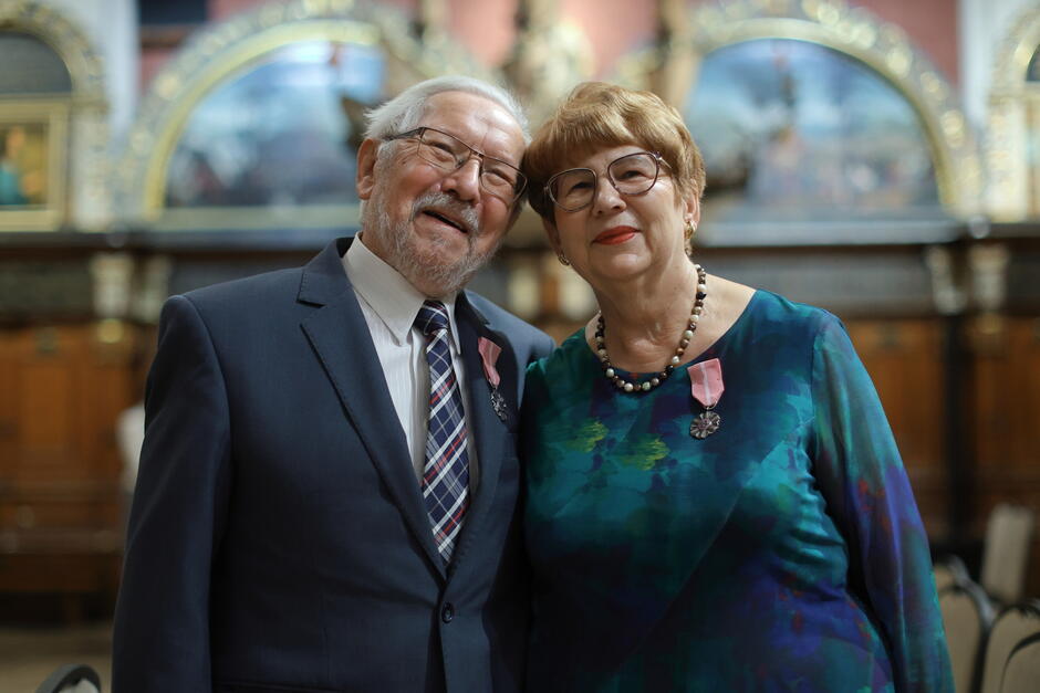 Na zdjęciu widzimy uśmiechniętą parę seniorów, stojącą blisko siebie, co podkreśla ich wzajemne ciepło i bliskość. Oboje są elegancko ubrani i odznaczeni medalami, co wskazuje na uroczysty charakter okazji, prawdopodobnie jubileusz małżeństwa