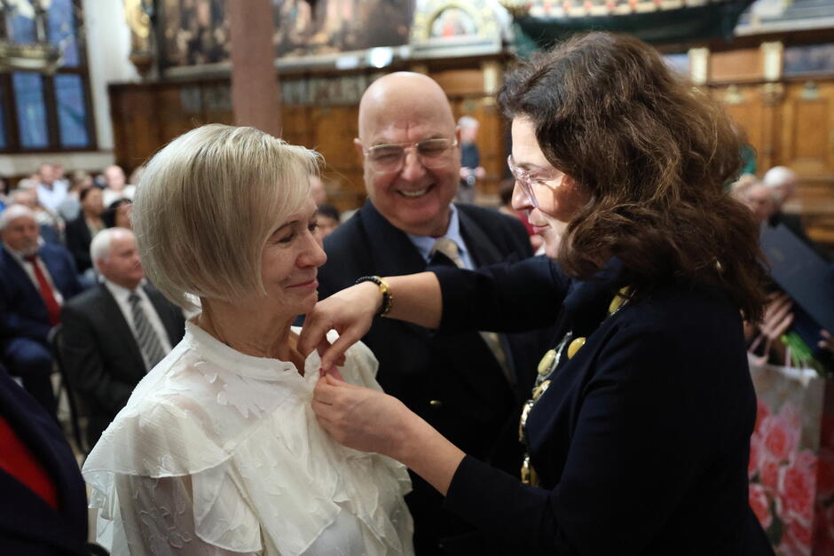 Na zdjęciu widzimy uroczystą chwilę przypinania odznaczenia kobiecie ubranej w białą elegancką bluzkę, która wygląda na poruszoną i uśmiechniętą. Obok niej stoi mężczyzna w garniturze z pogodnym wyrazem twarzy, a odznaczenie przypina kobieta w urzędowym stroju z łańcuchem, co podkreśla oficjalny charakter wydarzenia