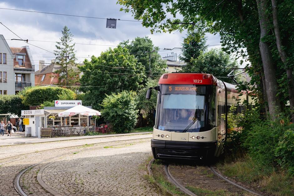 Na zdjęciu widzimy nowoczesny tramwaj linii numer 5, który porusza się po torach otoczonych zielenią, a w tle znajduje się przytulny bar typu Zapiekarnia  z ogródkiem. Scena rozgrywa się w miejskim krajobrazie z kamienicami i drzewami, tworząc spokojną atmosferę.