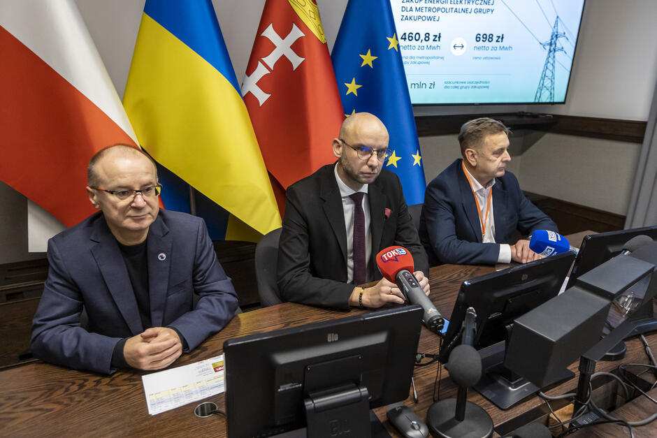 Na zdjęciu widzimy trzech mężczyzn siedzących przy stole konferencyjnym podczas oficjalnej konferencji prasowej. Na stole znajdują się mikrofony z logotypami mediów, takich jak TOK FM i inne, co wskazuje na obecność przedstawicieli mediów. W tle widoczne są flagi Polski, Ukrainy, Gdańska i Unii Europejskiej, co sugeruje, że wydarzenie ma charakter oficjalny i dotyczy współpracy międzynarodowej lub regionalnej. Na ekranie w tle znajduje się slajd z informacjami dotyczącymi zakupu energii elektrycznej, w tym cen netto za MWh oraz oszczędności dla grupy zakupowej, co sugeruje, że konferencja dotyczy kwestii energetycznych lub gospodarczych. Mężczyźni wyglądają na przedstawicieli władz lub organizacji zaangażowanych w zarządzanie takimi projektami. Atmosfera jest formalna, a uczestnicy są ubrani w eleganckie stroje, co podkreśla profesjonalny charakter spotkania.